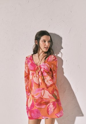 Frau mit langen dunklen Haaren, die ein orange-pinkes Blumenkleid trägt, steht vor einer strukturierten hellen Wand, hinter ihr fällt ein Schatten.