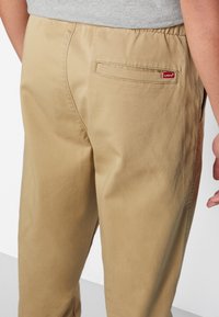 Beige Baumwollmix-Hosen mit elastischem Bund, einer aufgesetzten Gesäßtasche und einem roten Levi's-Logo-Anhänger in der Nähe der Tasche. Glatte Textur.