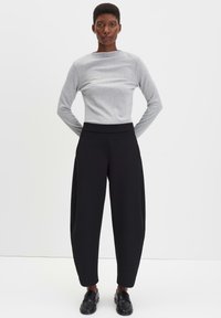Vrouw staat met handen achter de rug, gekleed in een lichtgrijze tops met lange mouwen, zwarte wijde broek en zwarte instappers, tegen een witte achtergrond.