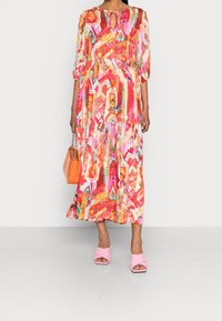 Maxikleid in leuchtendem Pink, Orange und bunten Mustern mit rundem Ausschnitt und Schlüssellochdetail. Kombiniert mit pinken Blockabsatzsandalen und einer orangefarbenen Tasche.
