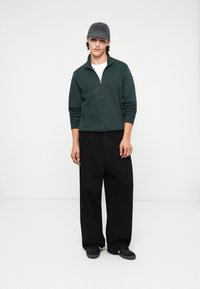 Donkergroen zip-up sweatshirt met de tekst "ORIGINALS", gecombineerd met zwarte wijde broek en een grijze baseballcap.