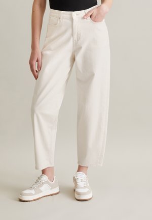 Partie inférieure du corps d'une personne portant un pantalon blanc ample taille haute et des baskets blanches, debout avec une main dans la poche.