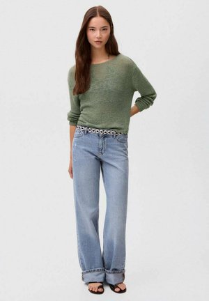 Junge Frau, die einen grünen Strickpullover und eine weite blaue Jeans trägt, steht selbstbewusst vor einem neutralen Hintergrund.