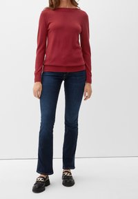 Rode gebreide trui met lange mouwen en een wijde halsopening, gecombineerd met blauwe flared jeans. De schoenen zijn zwarte loafers met een gouden accent.