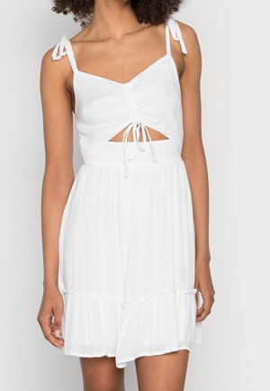 Femme portant une robe blanche sans manches avec des bretelles à nouer, un devant froncé, une découpe à la taille et une jupe à volants.