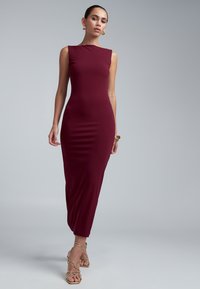 Woman wearing a sleeveless, fitted burgundy midi dress with gold strappy heels and matching gold earrings and bracelet against gray background.