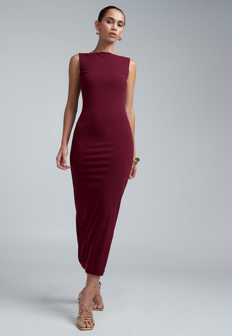 Woman wearing a sleeveless, fitted burgundy midi dress with gold strappy heels and matching gold earrings and bracelet against gray background.