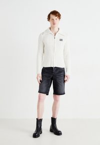 Chaqueta blanca de cremallera ribeteada con cuello, combinada con pantalones cortos de mezclilla negros y botas negras hasta el tobillo, de pie sobre un fondo claro.