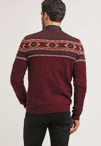 Pull en maille bordeaux avec un yoke à motif géométrique en vert, jaune et blanc. Poignets et ourlet côtelés.