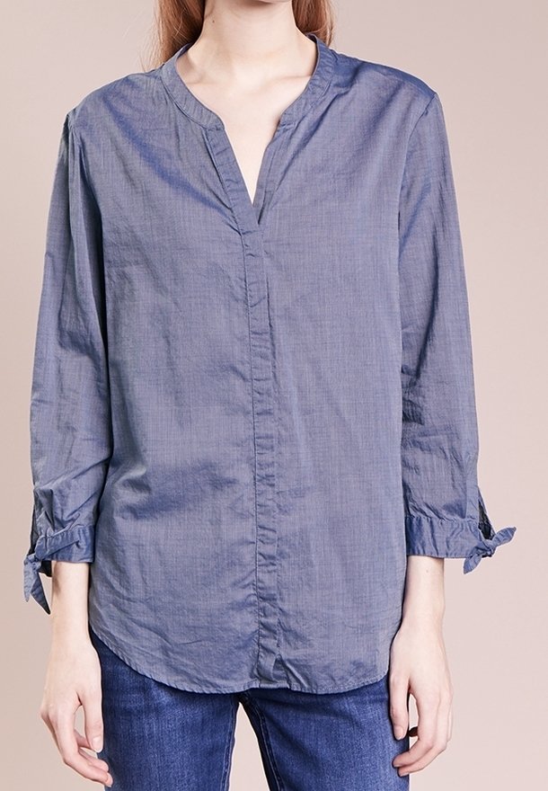 Femme portant un chemisier ample à manches longues bleu clair avec un col en V et des poignets noués, associé à un jean bleu.
