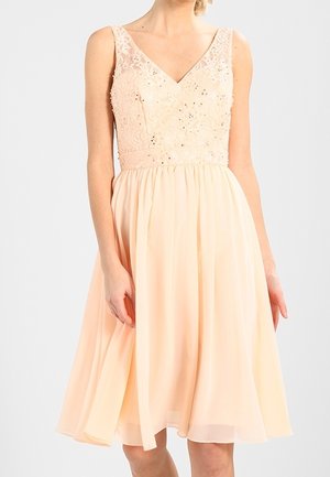 Bezrekawną brzoskwiniową sukienkę z koronkowym i cekinowym górnym elementem, dekoltem w kształcie litery V oraz falującą spódnicą do kolan nosi stojąca osoba.