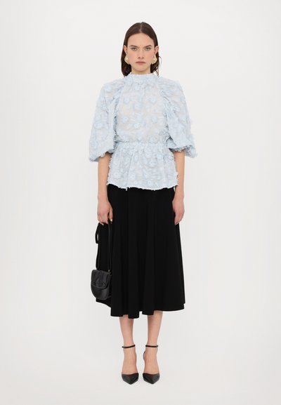 Bluză bleu deschis, transparentă, cu broderie florală și mâneci bufante, purtată cu o fustă neagră, până la genunchi. Modelul ține o geantă mică neagră.