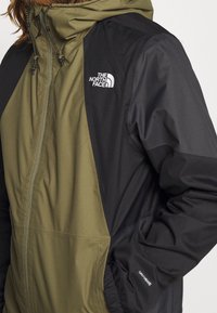 Chaqueta de hombre en verde oliva y negro con cierre de cremallera en la parte delantera, capucha y cordones ajustables. Presenta logos de la marca y puños elásticos.