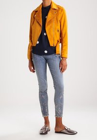 Gul mocka motorcykeljacka, marinblå skjorta med prickar, slitna ljusblå jeans med blommönster, och metalliska slip-on skor.