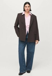 Blazer marrón de doble botonadura con raya diplomática, camisa rosa claro debajo, vaqueros azules de corte holgado y zapatos negros. Tela suave y diseño estructurado.