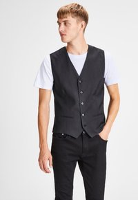 Chaleco negro con un diseño clásico, hecho de tela suave. Cuenta con cinco botones y dos bolsillos frontales. Lleva puesto sobre una camiseta blanca.