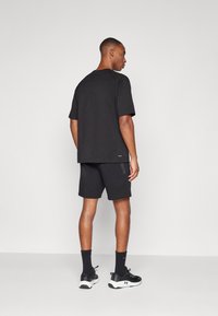 Homme debout, de dos, portant un t-shirt noir oversize, un short noir, des chaussettes noires et des chaussures de sport noir et blanc sur un fond clair uni.