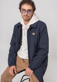 Marineblå lett jakke med trykknapper, med et lite fargerikt broderi på venstre bryst, brukt over en hvit hettegenser.