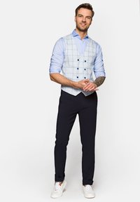 Jasnoniebieska kamizelka w kratkę z ciemnymi guzikami, noszona na jasnoniebieskiej koszuli, w zestawie z ciemnymi spodniami i białymi trampkami. Gładki, dopasowany fason.