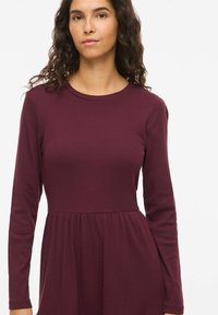 Robe bordeaux à manches longues avec une texture côtelée, un col rond et une jupe évasée. Tissu doux avec un corsage ajusté.