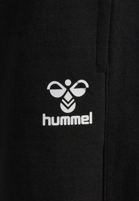 Białe stylizowane logo pszczoły z napisem „hummel” poniżej na czarnej tkaninie, wyśrodkowane w pobliżu pionowego szwu.