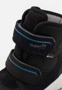 Černá vysoká teniska s suchým zipem, s texturovaným síťovaným svrškem, modrou akcentovou linií a logem GORE-TEX blízko podešve.