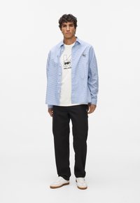 Modrá kockovaná košeľa cez biely tričko s potlačou, v kombinácii s čiernymi nohavicami a bielymi teniskami. Neformálny, voľný strih, s vyšitým detailom.