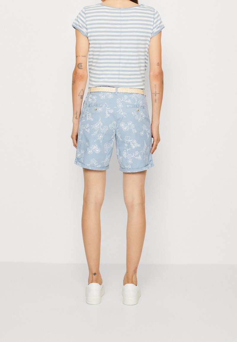 Personne debout portant un short fleuri bleu clair, une chemise rayée à manches courtes, des baskets blanches et une ceinture beige, vue de dos.