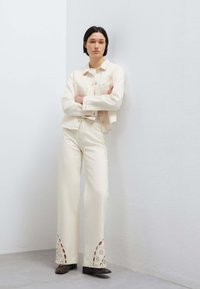 Frau mit kurzen dunklen Haaren steht an einer Eckwand und trägt eine cremefarbene Jacke und Hose mit ausgeschnittenen Blumendesigns an den Säumen sowie schwarze Schuhe.