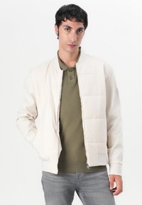 Helle beige Bomberjacke aus glattem Stoff, mit geripptem Kragen und Bündchen, ausgestattet mit einem Reißverschluss vorne und einer gesteppten Fläche auf der Brust.