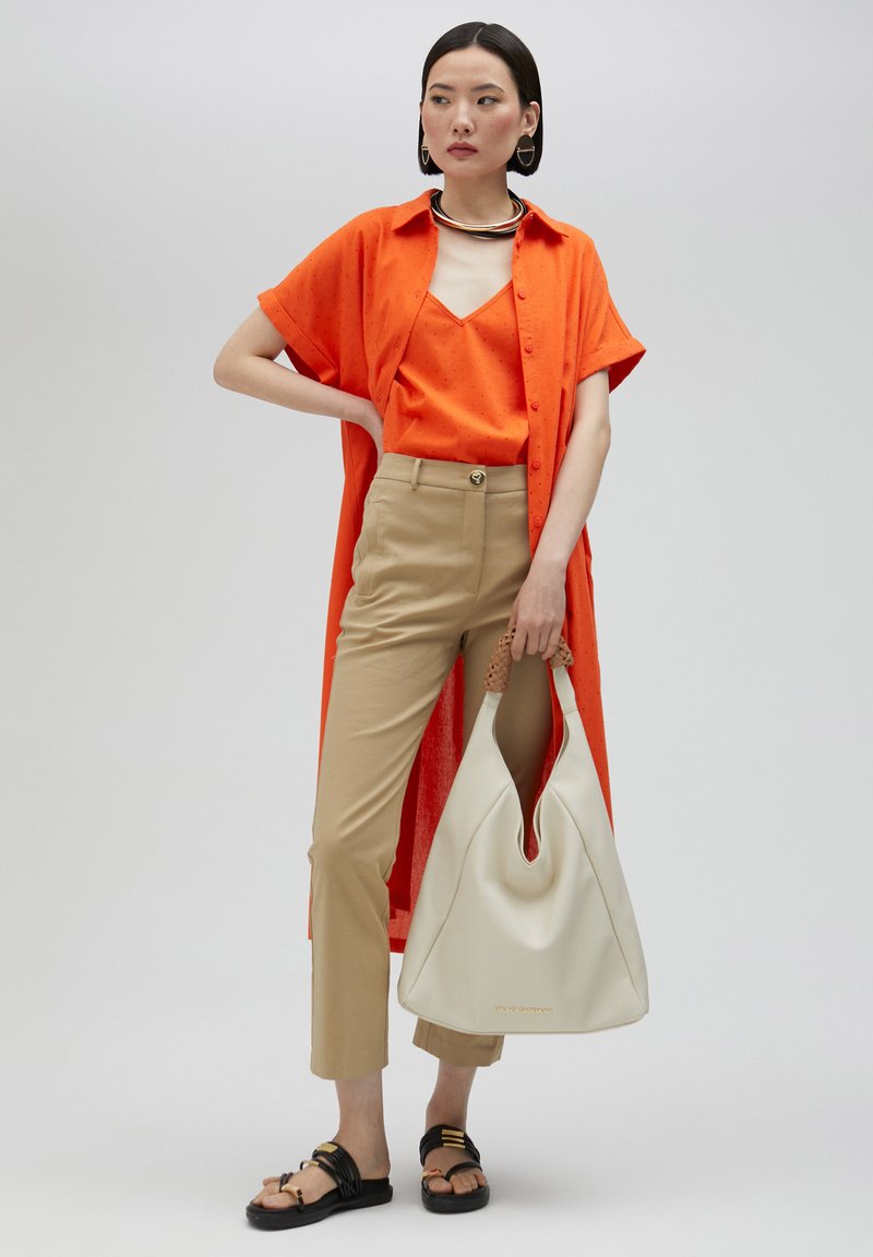 Le modèle porte une chemise orange vif et un débardeur superposé sur un pantalon beige ajusté. Elle tient un grand sac en cuir blanc et porte des sandales noires.