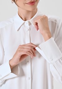 Camicia bianca con colletto a punta, bottoni in perlato e polsini con dettaglio a bottone e texture plissettata. Tessuto morbido.