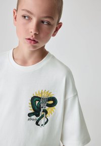 Baltas medvilninis marškinėlis su ryškiu grafiniu vaizdu – žalias gyvatė apvyniojęs skeletinę ranką, papuoštą geltonomis žaibo detalėmis.