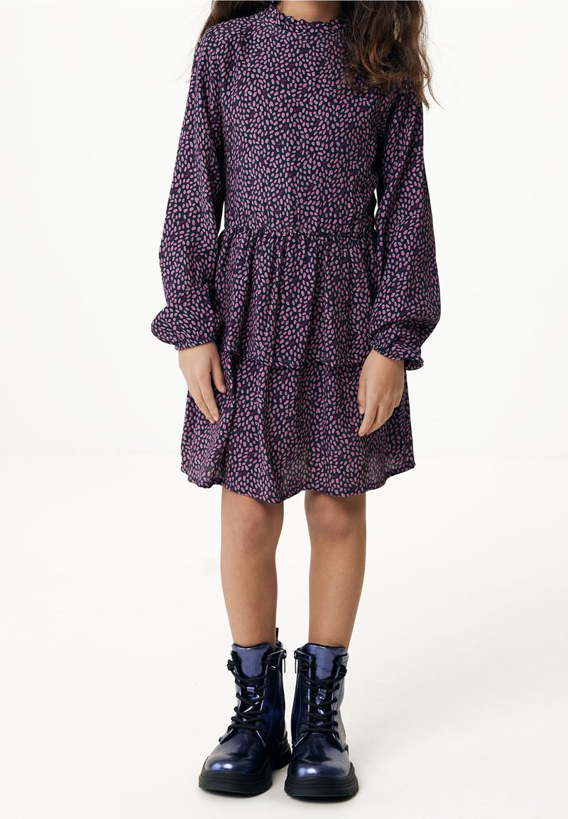 Enfant portant une robe violette à manches longues avec des taches roses et des bottes de combat noires brillantes, debout sur un fond blanc uni.