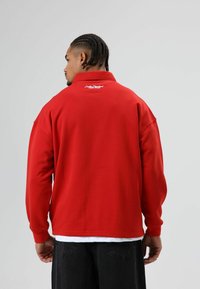 Rode trui met een hoge kraag en een geborduurd logo op de achterkant. Heeft een losse pasvorm en lange mouwen, gecombineerd met een donkere broek.