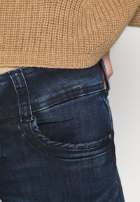 Jean en denim bleu foncé avec une texture lisse, présentant un design classique à cinq poches et de subtiles accents délavés au niveau de la poche arrière.