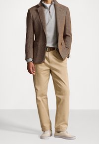 Hnědý herringbone blazer přes šedý svetr na zip, spárovaný se béžovými kalhotami a světlými teniskami, s hnědým opaskem.