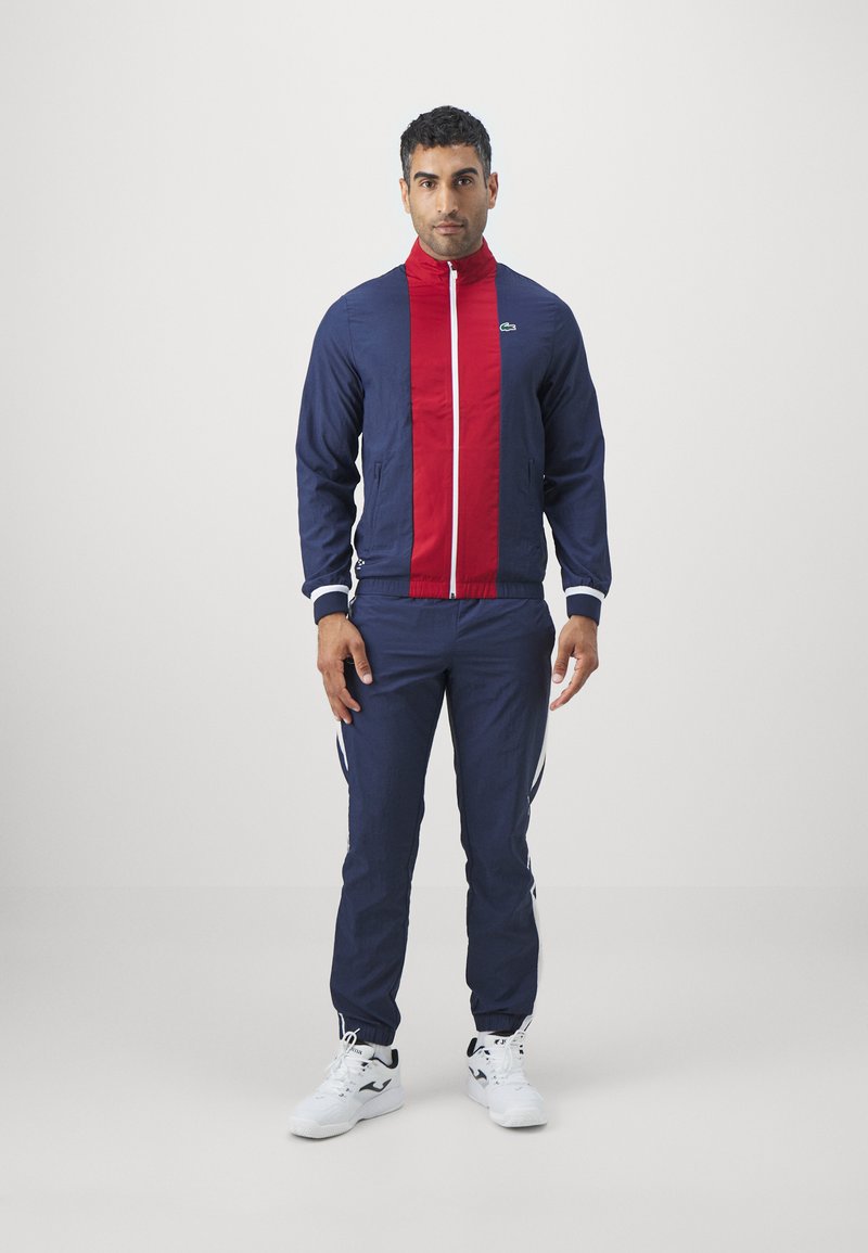 Survêtement bleu marine avec un panneau de fermeture éclair rouge, des accents blancs et des rayures sur les côtés. La veste a un col montant et des poches zippées. Baskets blanches.