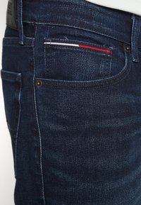 Jeans de mezclilla azul oscuro que presentan una vista en primer plano del bolsillo lateral con detalles de costura en rojo, blanco y azul, y acentos de remaches dorados.
