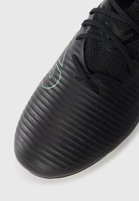 Čierne futbalové topánky s textúrovaným, ryhovaným zvrškom. Majú zelené logo a čierne šnúrky. Materiál vyzerá synteticky s hladkým povrchom.
