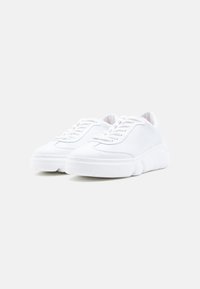 Ζευγάρι λευκά sneakers χαμηλού προφίλ με χοντρές σόλες και κορδόνια, σε απλό φωτεινό γκρι φόντο.