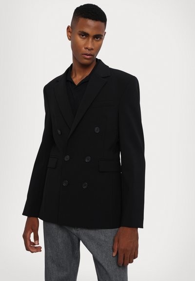 Blazer negru cu două rânduri de butoane, realizat dintr-un material neted, având rever înguste și buzunare laterale. Purta peste un top negru.