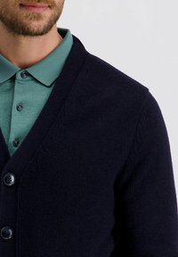 Marineblaue Strickjacke mit V-Ausschnitt, ausgestattet mit einem strukturierten Strickdesign, Vorderknöpfen und einem kontrastierenden grünen Hemd mit Kragen darunter.