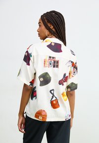 Kurzärmliges Hemd aus leichtem Stoff, mit einem cremefarbenen Hintergrund, der mit bunten Druckmotiven von Hüten, Lautsprechern und Accessoires verziert ist.