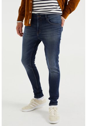Jeans ajustados de color azul oscuro con detalles desgastados, material de denim de algodón, diseño clásico de cinco bolsillos, combinados con zapatillas deportivas beige.