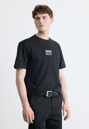 Mladý muž v čiernom tričku HUGO a čiernych nohaviciach s čiernym opaskom, jemne otočený do strany na jednoduchom pozadí.