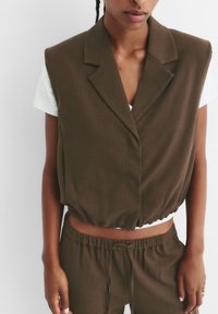 Gilet marrone con righe, dotato di colletto strutturato, in vita; abbinato a una maglietta grigia chiara e pantaloni marroni coordinati.