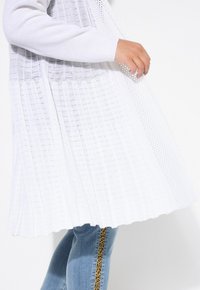 Vit stickad lång cardigan med en texturerad design och öppen vävstruktur, med ribbade ärmslut och en flödande silhuett.