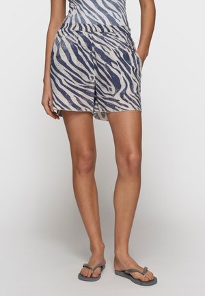 Ben iført beige og marineblå zebra-print shorts, én hånd i lommen, kombineret med grå klipklapper mod en ensfarvet baggrund.