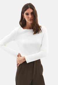 Kvinde med langt brunt hår iført hvid langærmet top og brune bukser, accessorized med guld hoops og ringe, neutral udtryk.